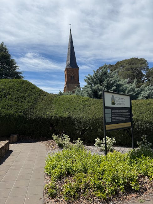 A well-maintained front garden scene outside St Johns Church Road in Canberra, featuring neatly trimmed green hedges along a paved pathway. In the foreground, there are low-growing flowering plants wi