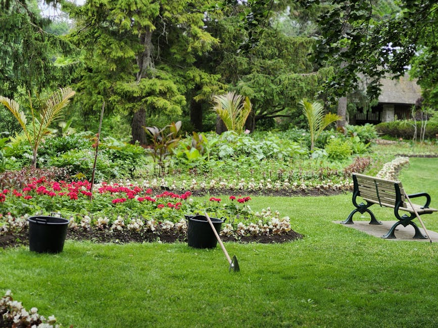 A neatly maintained garden featuring a lush, green lawn with freshly mowed grass, bordered by vibrant flower beds containing red, pink, and white blooms. In the background, mature trees and shrubs pro